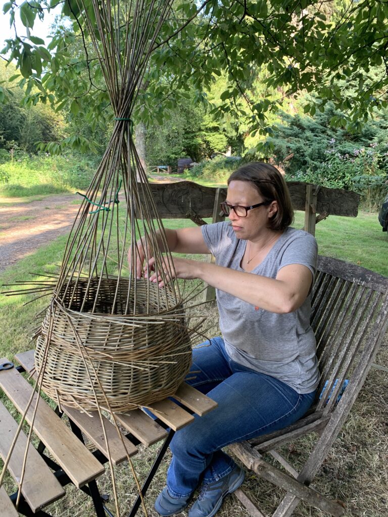 Lady sat in the Birtley Woodland Art Space weaving a basket on a traditional craft course with Hedges and Hurdles in a Woodland setting on the Birtley Estate