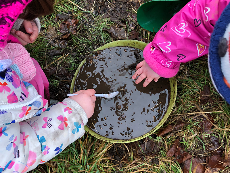 Hands of two children in pink coats dipping into a mud bath bucket. Surrey Hills Forest School activity